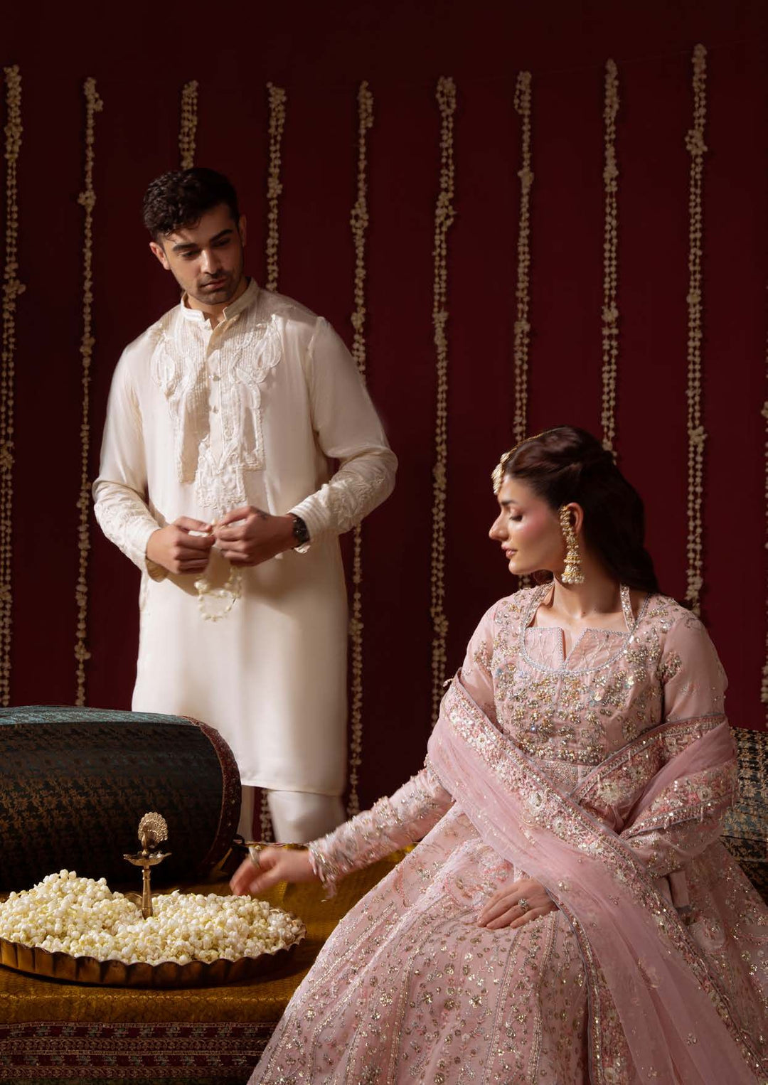 A couple seated on a divan in front of a deep red wall hung with flower garlands; the woman wears a blush-pink ensemble and reaches toward a bowl of flowers